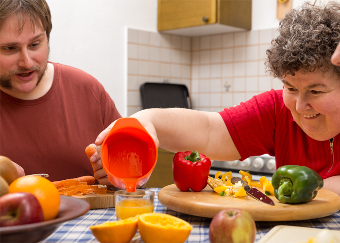 People sitting around a table and cooking. Chopped fruits and vegetables can be seen with one person pouring some liquid from an orange cup