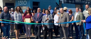 Several people are gathered around a man who is cutting a green and blue ribbon with oversized scissors for the grand opening of the Desert Hope Apartments