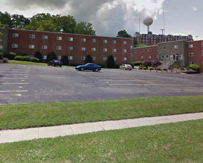 Exterior view of a multi-unit, three-story brick affordable housing complex with a parking lot and lush green trees under a clear sky