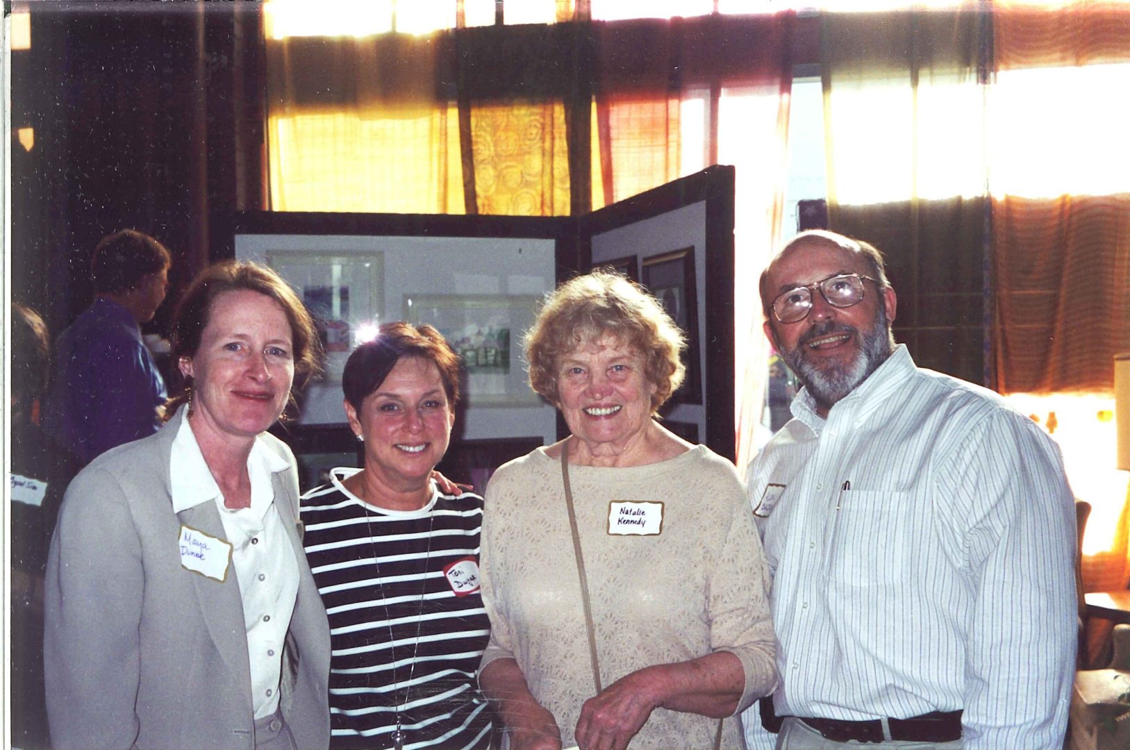 Group photo of four individuals at a social event. From left to right, a woman named Marla, a woman named Kim, a woman named Natalie Kennedy, and a man named Allen P. Baldwin. They are all smiling and looking towards the camera. The name tags on their clothing are visible, and a bright window forms the background.