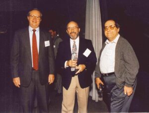 Allen P. Baldwin stands smiling in the center of a group of three men at a formal gathering. On his left is a man in a dark suit and red tie, and on his right is a man in a grey sweater and glasses. They all have name tags and appear to be engaged in conversation with drinks in hand.