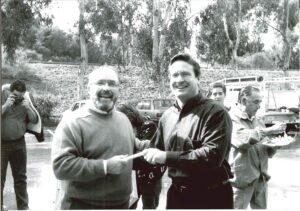 Outdoor candid shot of Allen P. Baldwin smiling and handing a piece of paper to another smiling man in a black jacket. In the background, various individuals are present, including one person taking a photograph and another person enjoying a meal.
