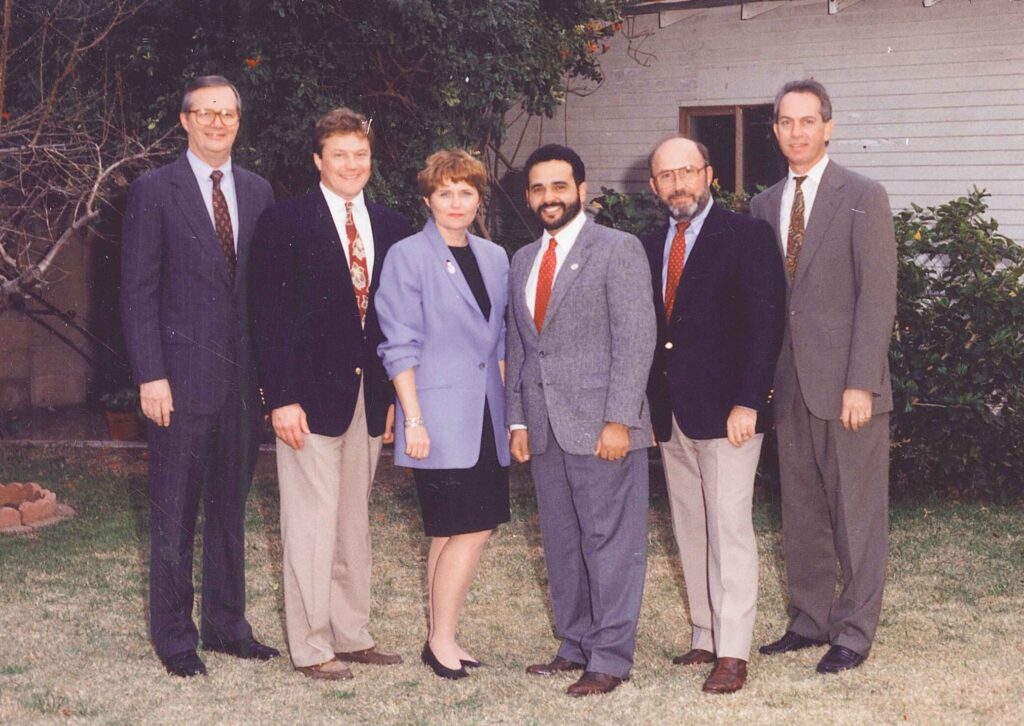 A formal group photo of six individuals standing in front of a house with a garden. From left to right, there are five men and one woman, all dressed in professional attire. Allen P. Baldwin is the fourth person from the left, wearing a suit with a red tie and glasses.