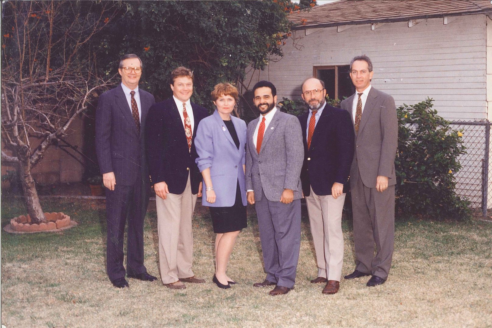 A formal group photo of six individuals standing in front of a house with a garden. From left to right, there are five men and one woman, all dressed in professional attire. Allen P. Baldwin is the fourth person from the left, wearing a suit with a red tie and glasses.