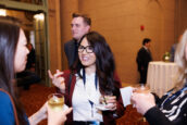 A woman with glasses talking animatedly, holding a drink, with others in conversation in the background at the Clearinghouse CDFI event.