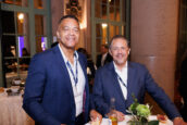 Two men smiling at the camera, both wearing business suits and event lanyards, seated at the Clearinghouse CDFI Annual Meeting.