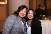 Two smiling women, one wearing a grey blazer and the other a blue blouse, standing together at the event, with other attendees in the background.