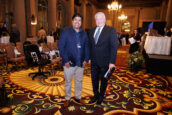 Two men standing and posing for the camera, one in a blue blazer and the other in a dark suit, both with conference ID tags, in a room with patterned carpeting at the Clearinghouse CDFI 2024 annual shareholders meeting.