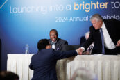A businessman in a suit is reaching over a table to shake hands with another person off-camera. They are at the front of a conference room with an audience and the "Launching into a brighter tomorrow" slogan on the screen behind.