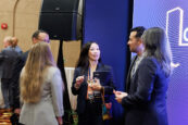 A businesswoman in a black blazer holding a glass and a brochure discusses with attendees in a lively networking session at the Clearinghouse CDFI event