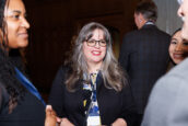 A businesswoman in a black blazer holding a glass and a brochure discusses with attendees in a lively networking session at the Clearinghouse CDFI event