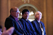 Three male attendees at the Clearinghouse CDFI's 2024 Annual Shareholders Meeting, appearing attentive and serious, with a chandelier visible in the softly lit background.