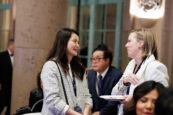 Two women engaging in a conversation, one smiling with a plate of hors d'oeuvres, at the Clearinghouse CDFI's 2024 Annual Shareholders Meeting, with other attendees in the background.