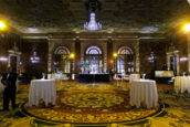 An elegant reception in the Gold Ballroom at the Biltmore in Los Angeles, set for the Clearinghouse CDFI's 2024 Annual Shareholders Meeting. The spacious room is lit with a warm glow highlighting the intricate ceiling details and patterned carpet. Round tables draped with white cloths are spread across the room, each adorned with a simple floral arrangement. A grand chandelier serves as a centerpiece above, and a stage with musical instruments is set up in the background, promising a night of entertainment