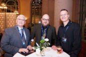 Three professionals smiling at the camera at a networking event, holding drinks with a conference setting in the background.