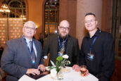 Three professionals smiling at the camera at a networking event, holding drinks with a conference setting in the background.