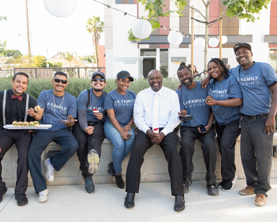 A group of cheerful L.A. Family Housing staff in branded T-shirts, enjoying a community event with food and smiles, showcasing team spirit and commitment.