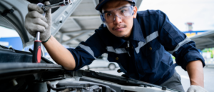 Focused auto mechanic in blue uniform inspecting and repairing a car engine, using a screwdriver in a well-equipped garage.